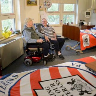 Two elderly individuals sit in a room decorated with banners, engaging in conversation.