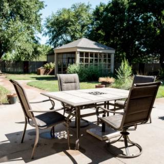 A backyard patio with a table and chairs set, surrounded by lush greenery and a gazebo in the background.
