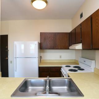 A compact apartment kitchen with wooden cabinets, a white refrigerator, stove, and a double sink.