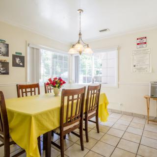 Bright dining room with a yellow tablecloth and wooden chairs, featuring a cozy chandelier and natural light from large windows.