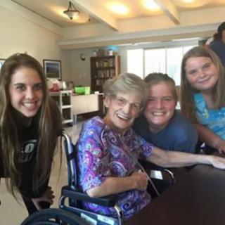 A happy group gathered around an elderly woman in a wheelchair indoors.