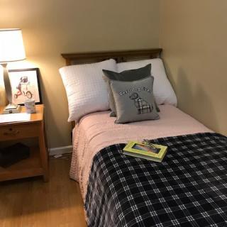 A cozy bedroom with a single bed covered in pink and black bedding, accompanied by a nightstand with a lamp and book.