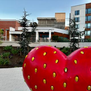 A vibrant strawberry heart sculpture in the foreground, with modern buildings and greenery in the background.