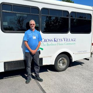 A man stands next to a community bus from Cross Keys Village, promoting local transportation services.