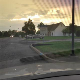 A tranquil sunset scene in an urban area, featuring a road lined with trees and parked vehicles.