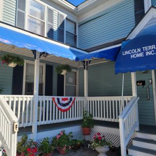 A quaint house with a porch featuring an awning and patriotic flag decorations, surrounded by potted flowers.