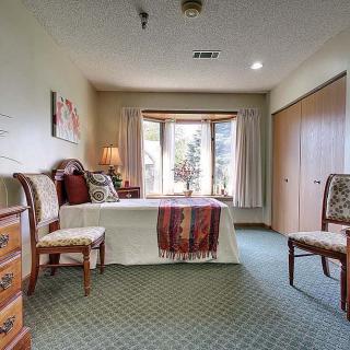 A cozy bedroom featuring a bed with decorative pillows, chairs, a dresser, and large windows providing natural light.