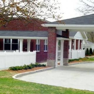 A brick building with a white fence and driveway surrounded by grass and trees.