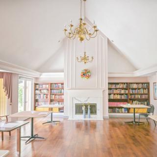 A spacious library with a high ceiling, featuring a central fireplace, elegant chandelier, and bookshelves filled with books.
