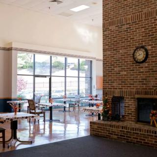 A spacious cafeteria with tables and chairs, featuring a brick fireplace and large windows.