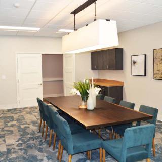 A modern meeting room featuring a long conference table surrounded by teal chairs, illuminated by a stylish overhead light fixture. The room includes wall-mounted cabinets and decorative artwork.