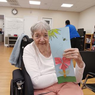 An elderly woman proudly displaying her colorful craft creation featuring a flower.