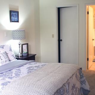 A cozy bedroom featuring a neatly made bed, a lamp on the nightstand, and an open doorway leading to another room.