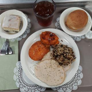 A meal served on a tray featuring turkey slices, wild rice, cooked carrots, a small sandwich, bread roll, and a drink.