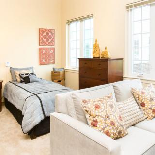 A bright and cozy bedroom with a neatly made bed and a comfortable sofa, complemented by stylish decor and ample natural light from large windows.