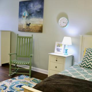 A cozy bedroom featuring a green rocking chair, a nightstand with a lamp, and artwork on the wall.