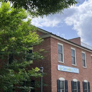 A brick building with green shutters and a sign, surrounded by trees under a partly cloudy sky.
