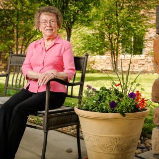 An elderly woman smiling while sitting on a bench in a garden during the daytime.