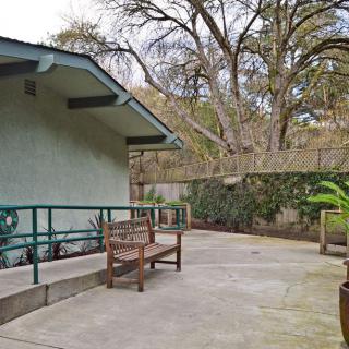 A serene patio area with a wooden bench, adjacent to a house surrounded by trees and landscaping.