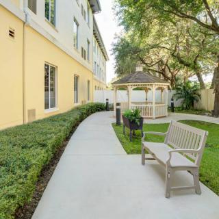 A serene garden area with a bench, pathway, and gazebo surrounded by lush greenery next to a building.