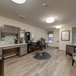 A modern salon interior featuring comfortable chairs, a styling station, and a kitchenette with wood flooring.