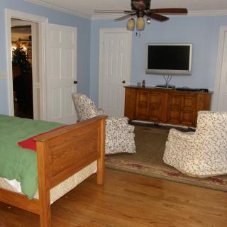 A cozy bedroom with a wooden bed, two chairs facing a television, and a ceiling fan.
