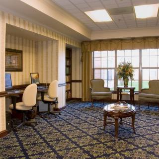 A cozy room with computers, desks, and chairs, featuring a carpeted floor and large windows with curtains.