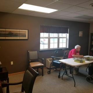 An elderly person sits at a table in a bright room, reading by the window.