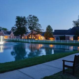 A peaceful evening scene with houses reflecting on a pond, illuminated by soft lighting and surrounded by trees and a pathway.