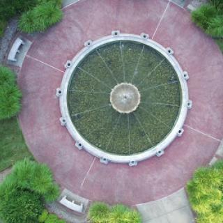 Aerial view of a circular fountain surrounded by lush greenery and benches in a park.