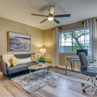 A cozy living room doubles as a home office, featuring a sofa, desk, and large window with natural light.