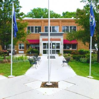 A brick building entrance with a pathway lined by flags, surrounded by trees and outdoor seating.