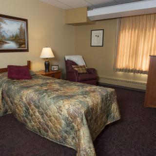 A cozy bedroom with a single bed, lamp, and armchair, featuring warm lighting and decor.