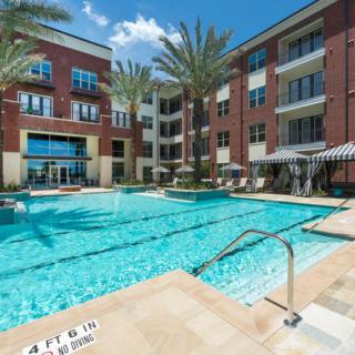 A modern apartment complex with a swimming pool surrounded by palm trees on a sunny day.