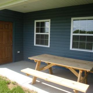 A patio area with a wooden picnic table outside a house featuring two windows.