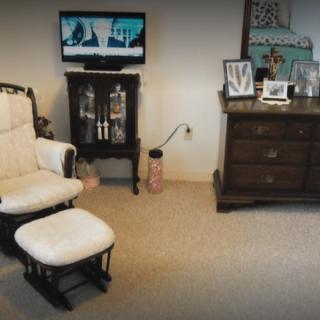A cozy living room with a rocking chair, ottoman, TV on a stand, and a dresser displaying framed photos.