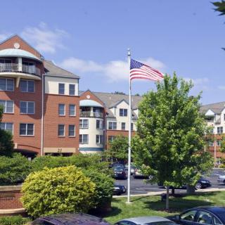 A landscaped area with a multi-story building, an American flag, and parked cars under a partly cloudy sky.