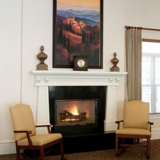 A cozy living room with a fireplace, flanked by two chairs and adorned with a landscape painting above the mantel.