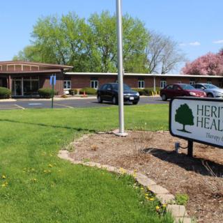A healthcare facility with a sign in the foreground, surrounded by a parking lot and greenery.