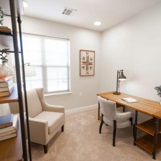 A cozy home office with a wooden desk, upholstered chairs, and a bookshelf by the window.