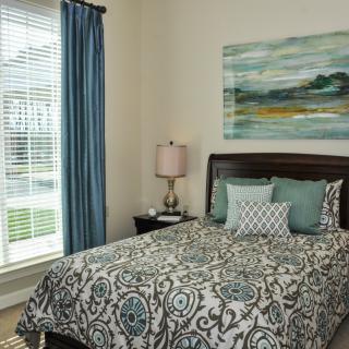 A cozy bedroom with patterned bedding, decorative pillows, and a painting above the wooden headboard. A lamp sits on the nightstand beside a window with blue curtains.