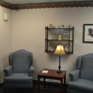 A cozy waiting room with two upholstered chairs, a lamp on a side table, and a shelf displaying decorative items.