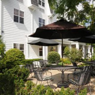 A charming patio area with tables and chairs under large umbrellas, surrounded by lush greenery and adjacent to a white building.