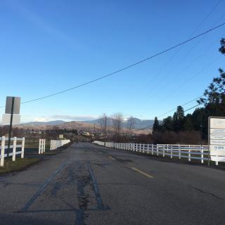 A quiet road flanked by white fences, with trees lining the roadside and mountains in the distance under a clear blue sky.