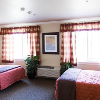A hotel room with two beds, decorated with patterned curtains and a framed painting between large windows.