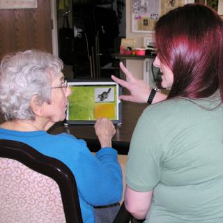 An elderly person engages with a caregiver, exploring content on a tablet together.