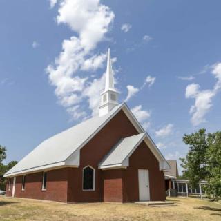 A traditional brick church with a prominent steeple stands under a partly cloudy blue sky.