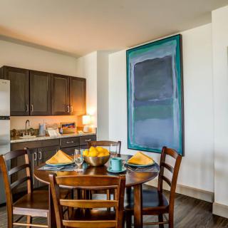 A modern dining area with a wooden table set for four, adjacent to a compact kitchen, featuring contemporary artwork on the wall.
