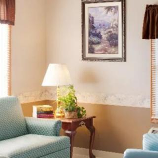 A cozy living room corner featuring a comfortable chair, side table with a lamp and books, and a decorative painting on the wall.