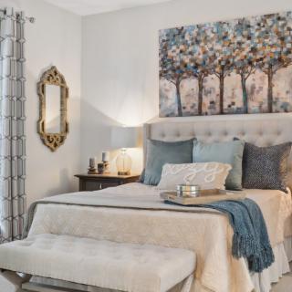 A cozy and elegantly designed bedroom featuring a bed with multiple pillows, a soft blanket, an artistic wall piece above the headboard, a decorative mirror, and a nightstand with a lamp.
